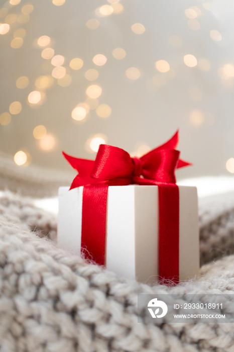 Christmas gift box with red bow on warm knitted sweater and defocused lights on background