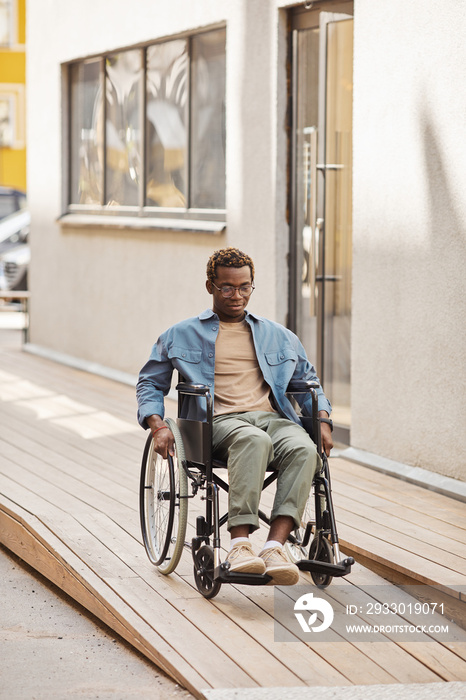 Disabled young African-American man using manual wheelchair while moving down ramp by house