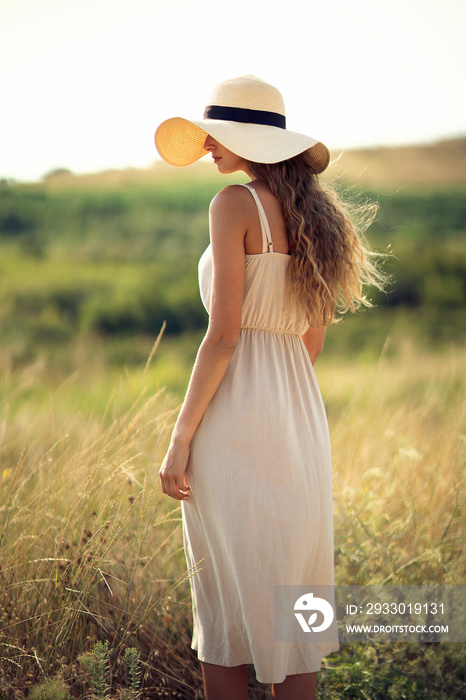 Back view of beautiful girl who walks on field while wearing a sunhat and midi dress. Lifestyle