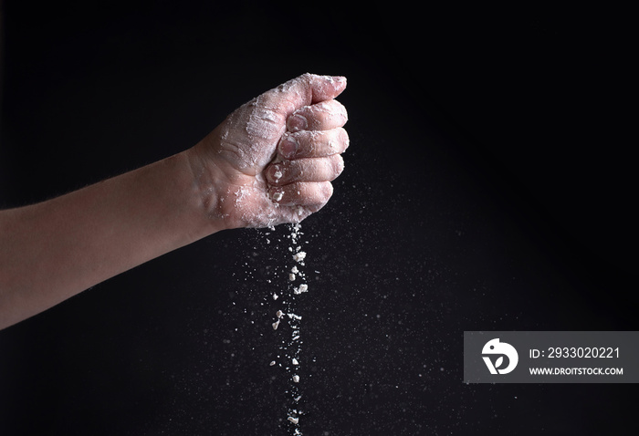 Female hand pouring flour on a black background, flying white powder.