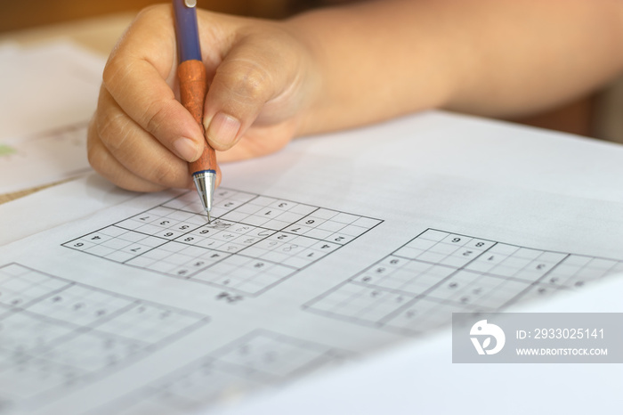 Solve sudoku puzzle with pencil as hobby by senior woman on wooden office desk. Player insert numbers into grid consisting of nine squares subdivided into further nine smaller squares.