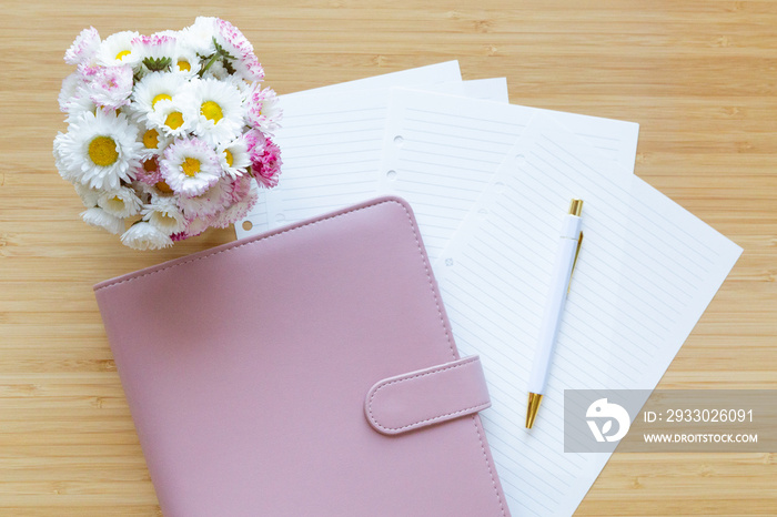 Flat lay, top view of a pastel pink planner, flower bouquet and stationery on a wooden desk.