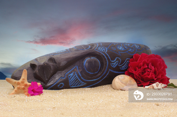 Wooden Tiki Mask on Sand With Flowers and Blue Sky Background