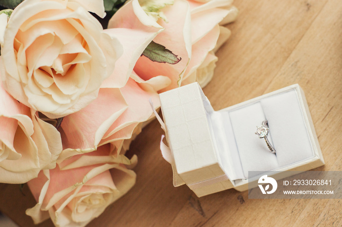 Close up of engagement ring in gift box with rose flowers on wooden table. Marriage proposal concept by means of flower bouquet and gift. Wedding proposal for beloved woman