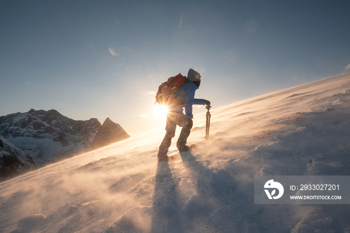 Man traveler with tripod are climbing on slope hill on Ryten mount in blizzard at sunset