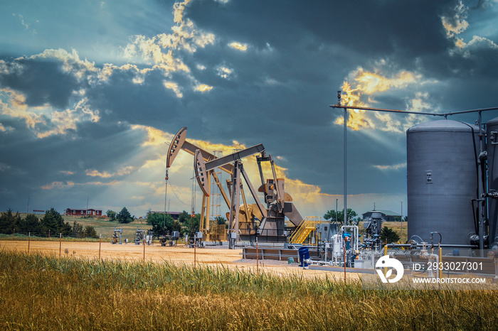 Two pump jacks and tanks and oil field equipment in fenced area in pasture under dramatic skies with houses on hill on horizon - summer