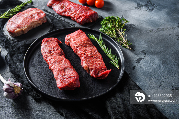 Raw denver, top blade,  tri tip steak on a black plate and stone slate with seasonings, herbs  grey concrete background. Side view close up selective focus new wide angle space for text