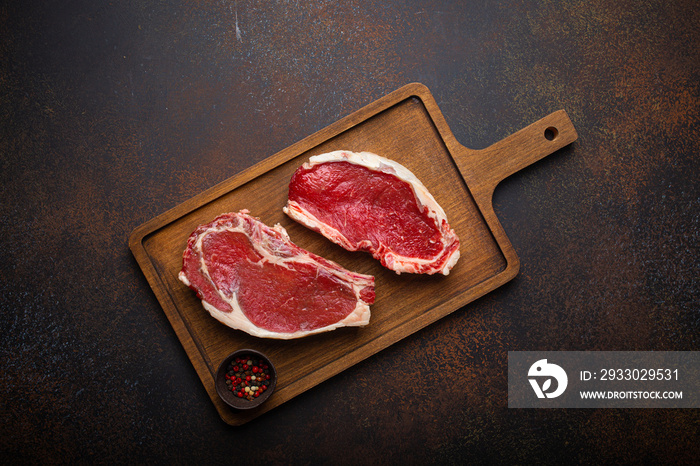 Two raw uncooked meat beef rib eye marbled steaks on wooden cutting board with seasonings on dark rustic background ready to be grilled from above, preparing dinner with meat