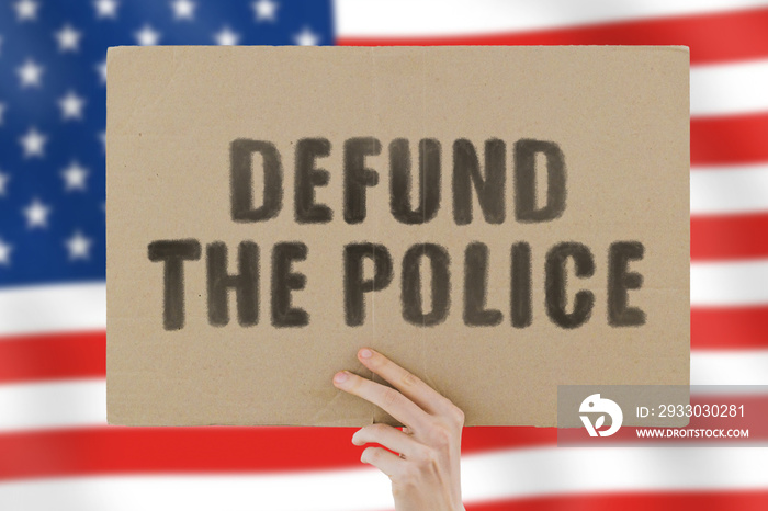 The phrase   Defund the police   on a banner in men’s hand with blurred American flag on the background. Violence against black. Fight. Police reform. Occupation. Job. Protest. Brutality. Equality