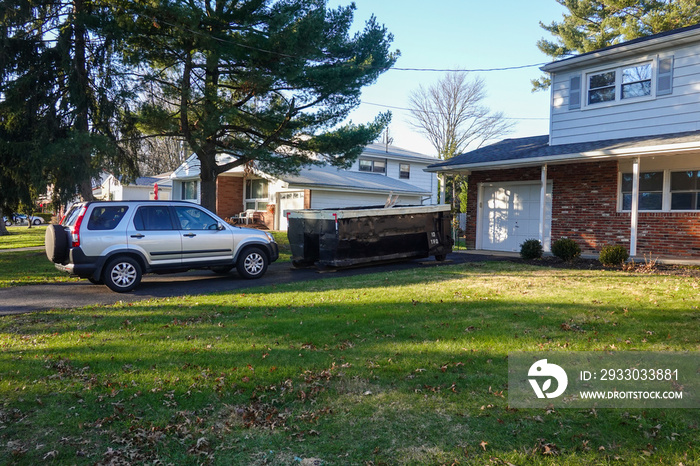 A small short beat up black and white dumpster containing wood debris located in a driveway in front of a garage door