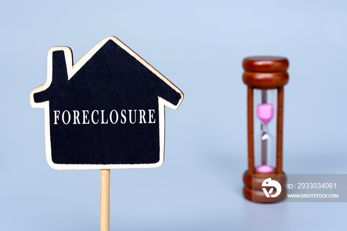 Foreclosure text on wooden house model with blurred hourglass background.