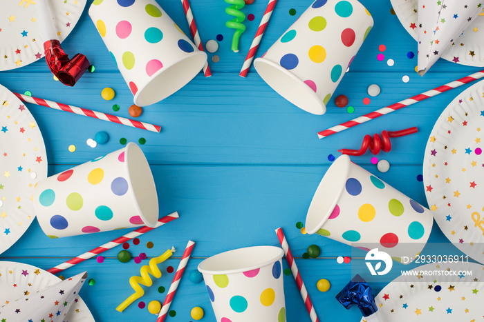 Top view photo of party table decoration paper plates cups hats pipes straws spiral candles sequins and confetti on isolated blue wooden table background with blank space