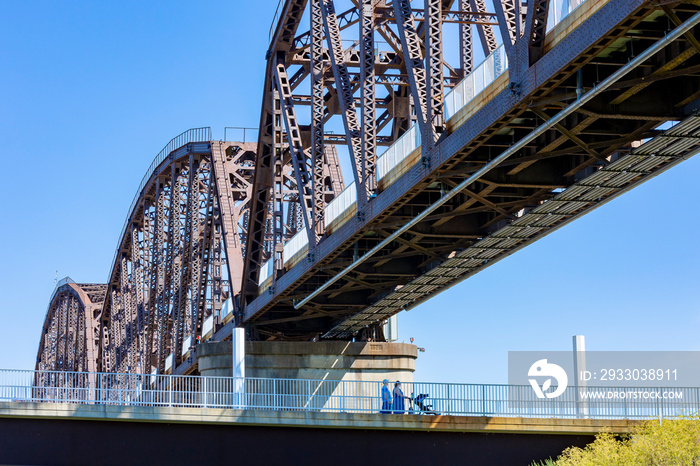 Two women and a baby buggy walking on a pedestrian ramp leading to an old, former railroad bridge that is now a pedestrian bridge.