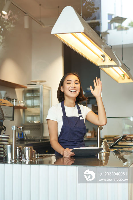 Vertical shot of happy young barista, asian cafe worker waves at client, receives orders behind counter, using tablet as POS terminal, working in coffee shop