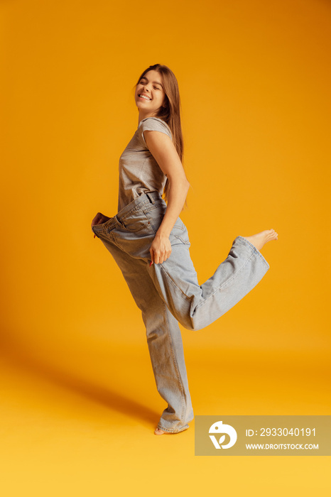 Portrait of young cheerful woman in big size jeans isolated over yellow background. Weight loss concept.