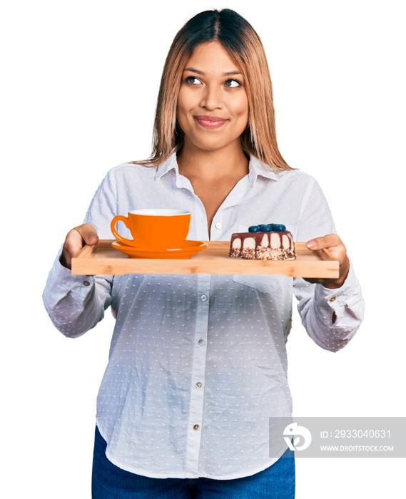 Young hispanic woman holding tray with coffee and cake smiling looking to the side and staring away thinking.