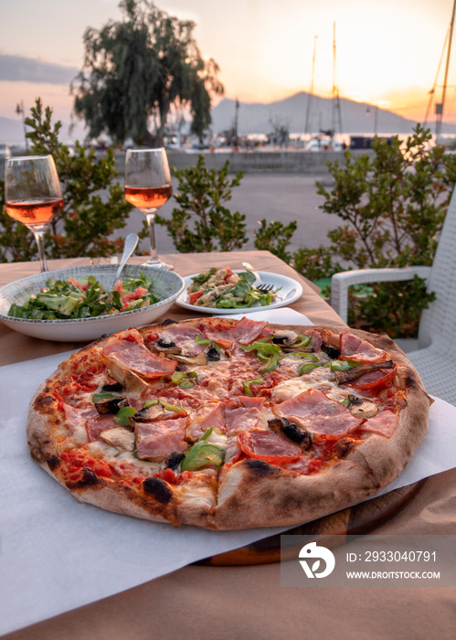 Delicious pizza with cheese, bacon, salami, mushrooms and green pepper and two glasses of wine against the background of the port in Greece