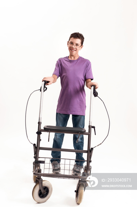 Full length shot of teenaged disabled boy with cerebral palsy in the glasses smiling at camera, taking steps with his walker isolated over white background