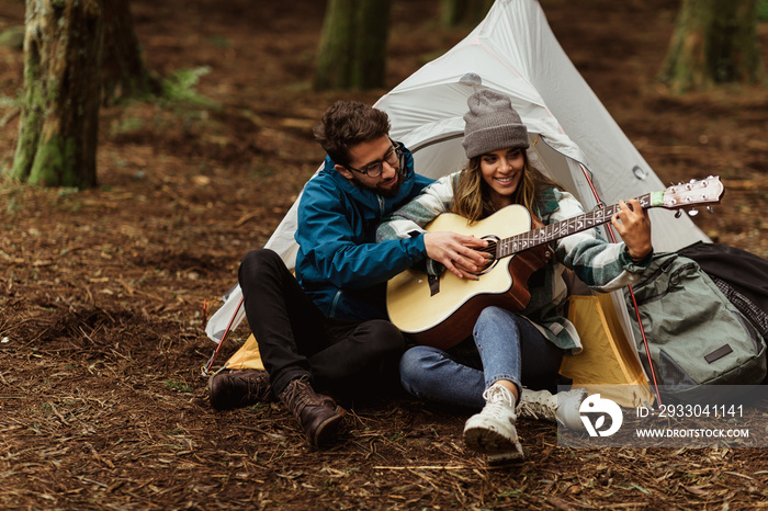 Cheerful millennial caucasian husband and wife in jackets sit in cold forest, enjoy adventure, do lesson, play guitar