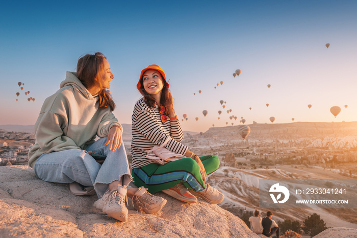 Girl friends sitting on a clifftop viewpoint and admiring view of majestic flying hot air balloons in Cappadocia