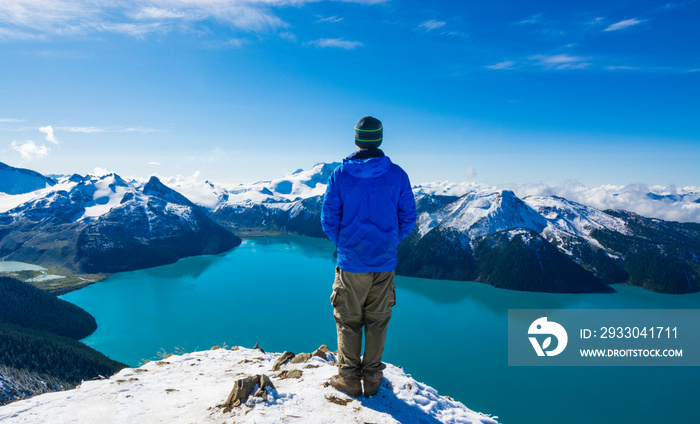 Adventurous athletic male hiker standing at the summit of Panorama Ridge, looking at Garibaldi Lake on a beautiful sunny day.