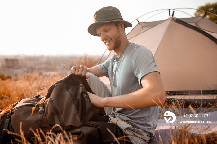 A man in a hat packing his backpack while sitting near the tent