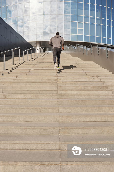 Low angle view of anonymous Black male athlete running up concrete steps towards contemporary office building while having outdoor workout