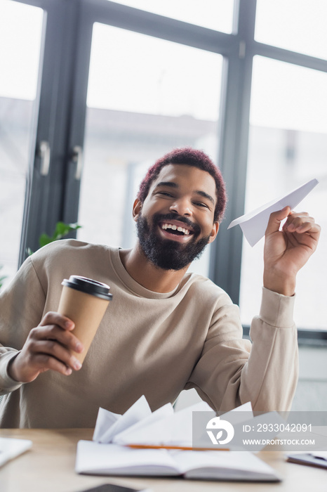 Happy african american businessman holding paper plane and takeaway coffee in office