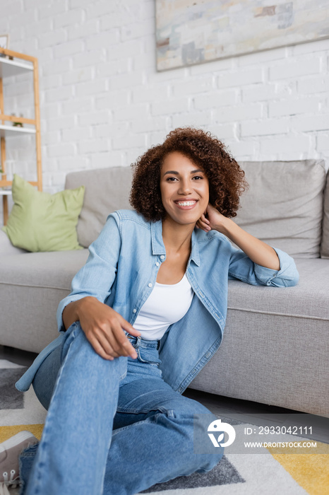 smiling african american woman with curly hair sitting near modern sofa in living room.