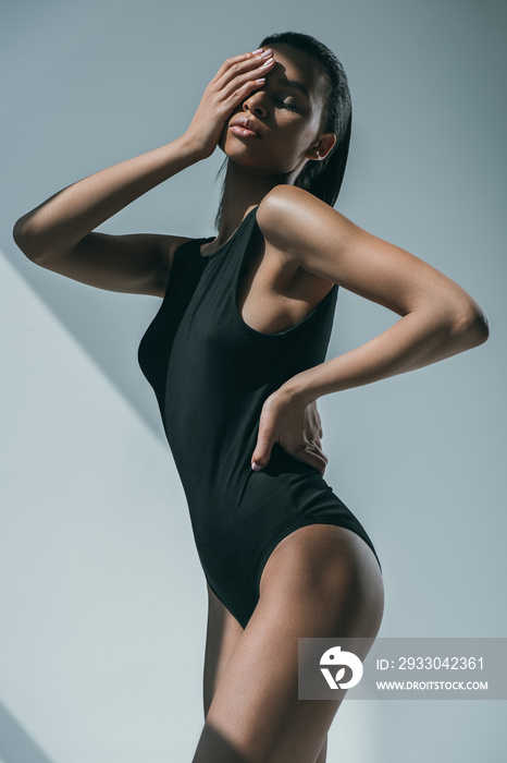 african american trendy woman with slim body touching her face and posing in black bodysuit