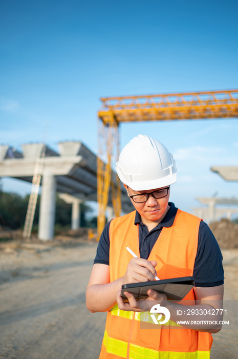 Smart Asian worker man or male civil engineer with protective safety helmet and reflective vest using digital tablet for project planning and checking architectural drawing at construction site.