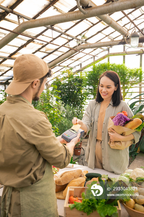 Pretty young Asian woman in casual outfit holding paper bag of organic products and making contactless payment in grocery store