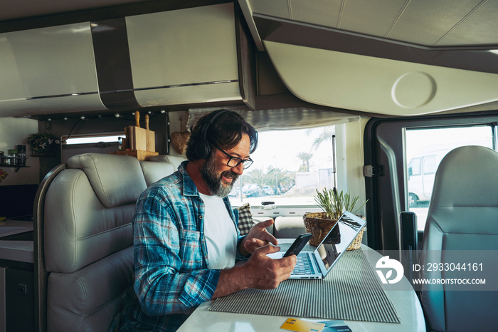 Traveler modern online people job business lifestyle. One happy mature man using phone and computer inside a camper van motorhome vehicle. Vacation travel and roaming connection technology worker