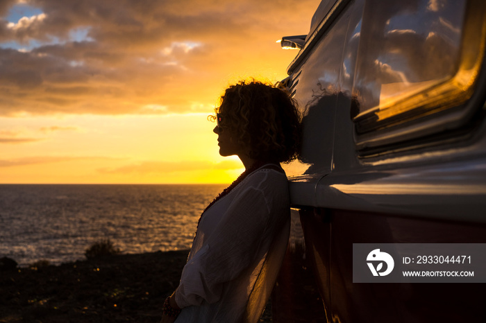Travel and wanderlust lifestyle destination concept with beautiful woman in silhouette and red vintage van enjoying the coloured orange sunset in front of the ocean view - independence and people