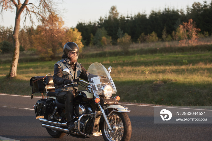 Bearded biker in sunglasses, helmet and black leather clothing riding cruiser motorcycle along empty asphalt country road by beautiful tall trees with golden foliage on sunny autumn evening.