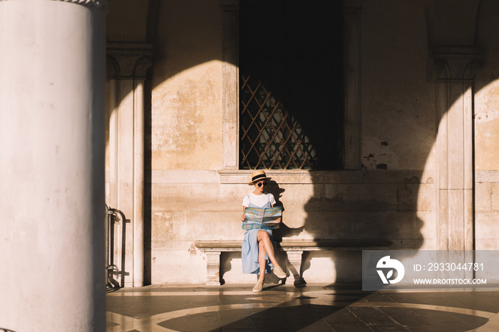 Focused young woman in hat holding map and sitting on bench at building