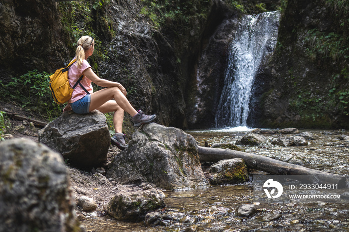 Woman resting during hike in wonderful nature. Hiker sitting on rock near stream with waterfall in forest at natural parkland Mala Fatra, Slovakia