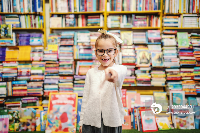 Adorable smiling little caucasian girl with eyeglasses and ponytail pointing with sword toy and looking at camera while standing in bookstore.