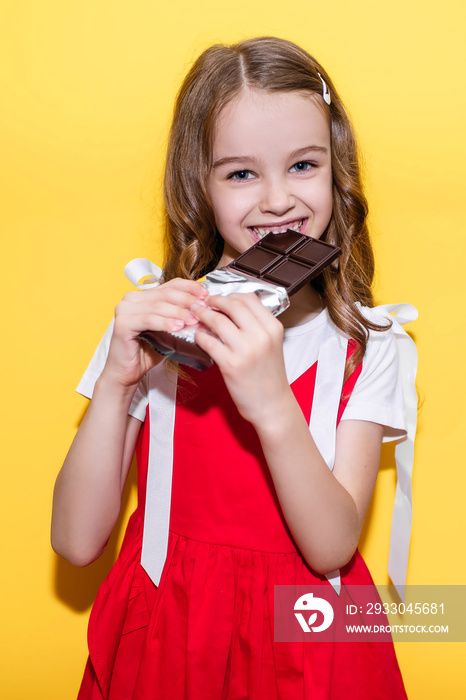 Little girl bites a bar of chocolate on a yellow background. Happy child with face and hands covered in chocolate. The concept of children’s happiness and love for sweets
