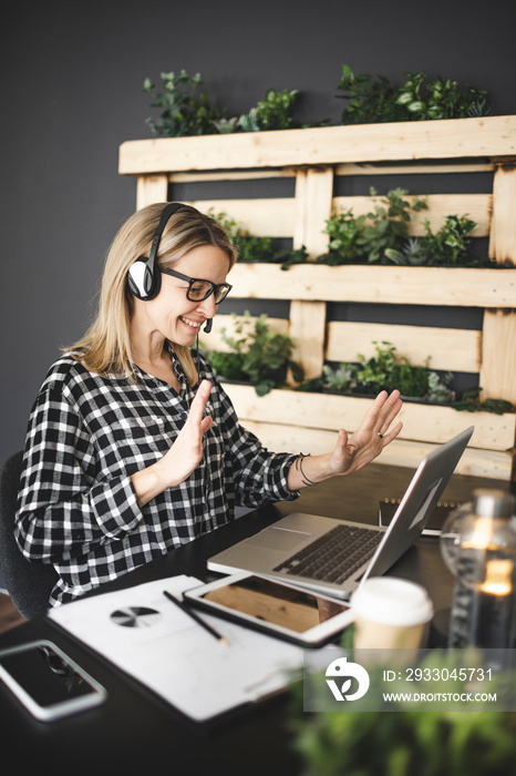 pretty, young blond businesswoman with black and white checked shirt is sitting in a sustainable, ecological office with a headset and is having a web meeting on her laptop