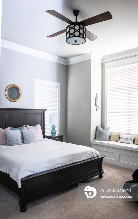 A light and bright gray master bedroom with a window seat, bed and a fan