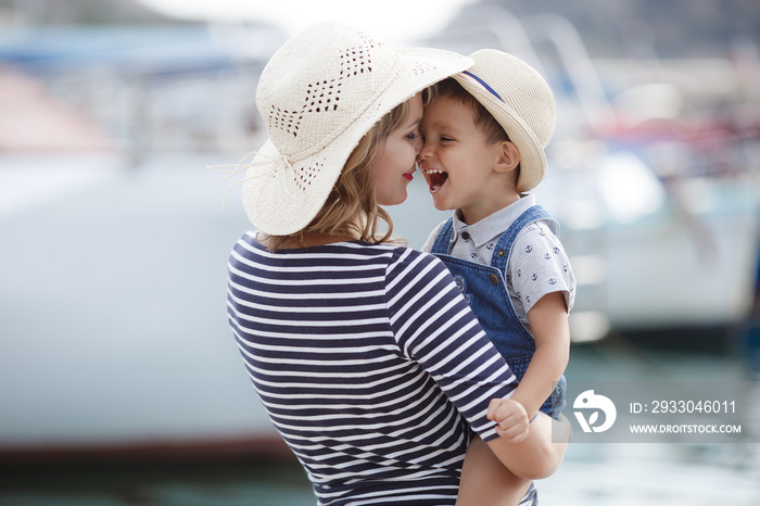 Mother and little son spend time outdoors in the summer together, sitting on a pier near boats and boats.Young mother plays with her baby on the promenade.Summer vacation for two by the sea. Mother an