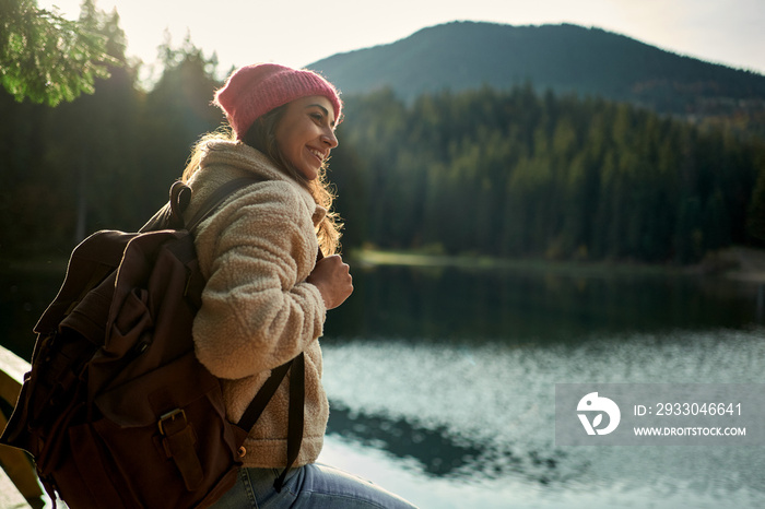 Side view backpacker smiling woman admiring mountain lake among forest with sunset light around