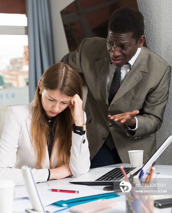 Frustrated woman sitting at office desk with disgruntled boss behind