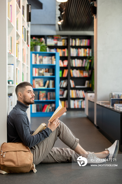 Young distracted man with open book looking aside while sitting on he floor against bookshelf