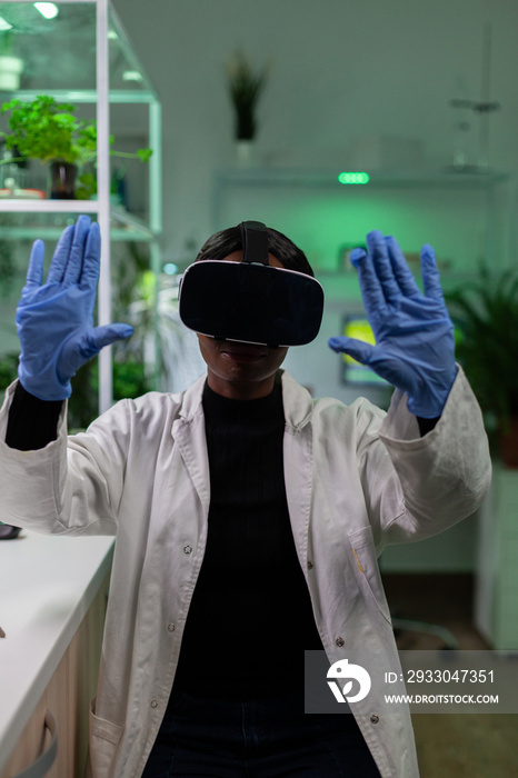 African american chemist woman wearing virtual reality headphones during microbiology experiment working in biological hospital laboratory. Scientist doctor analyzing genetically modified plants