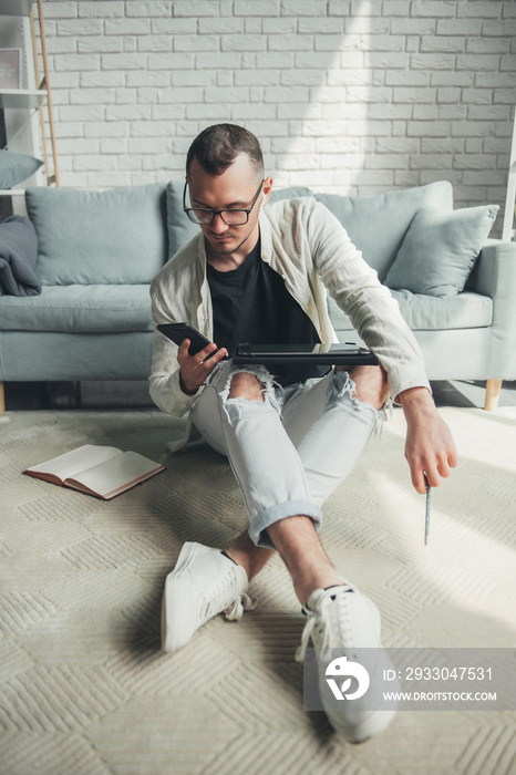 Caucasian man sitting on the floor near the sofa at home using a digital tablet, mobile phone for searching information in internet. Office, university library