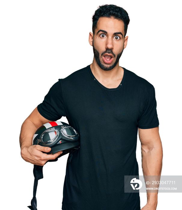 Hispanic man with beard holding motorcycle helmet scared and amazed with open mouth for surprise, disbelief face