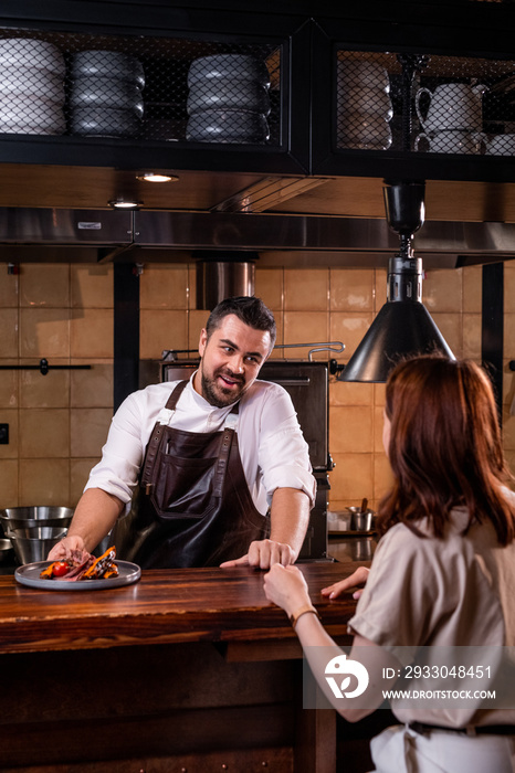 Handsome restaurant chef in apron standing at counter in open kitchen and telling about dish to guest