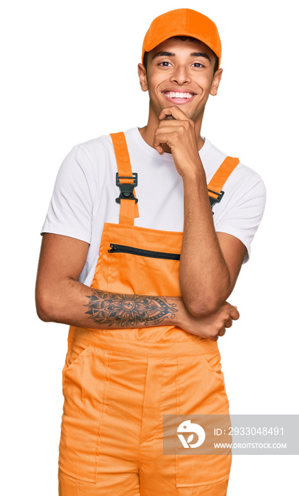 Young handsome african american man wearing handyman uniform looking confident at the camera with smile with crossed arms and hand raised on chin. thinking positive.